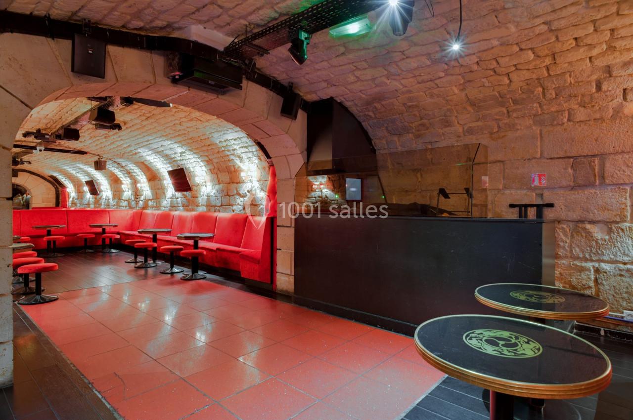 Salle voûtée en pierre avec banquettes rouges, tables basses et comptoir, éclairage tamisé.