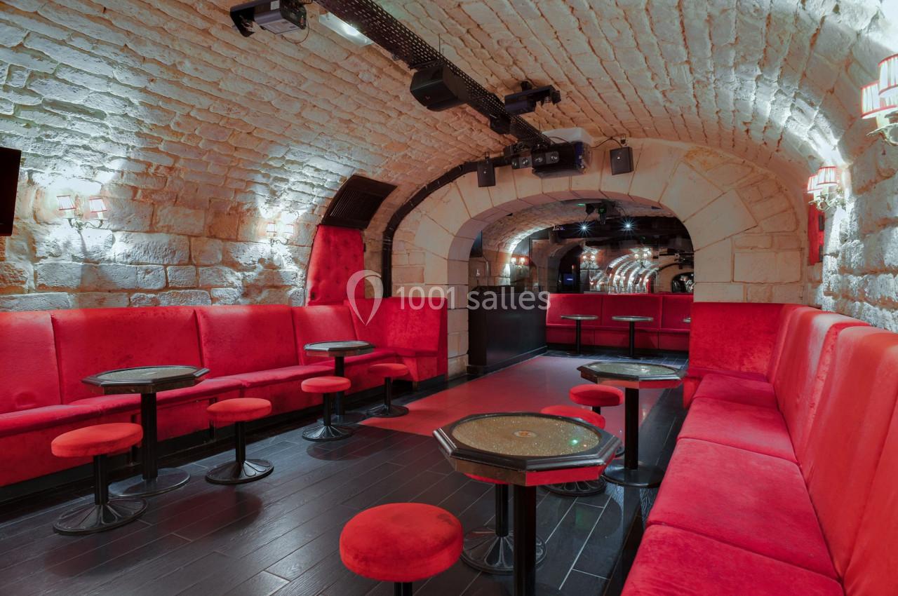 Salle voûtée en pierre avec banquettes rouges, tables basses et éclairage tamisé.