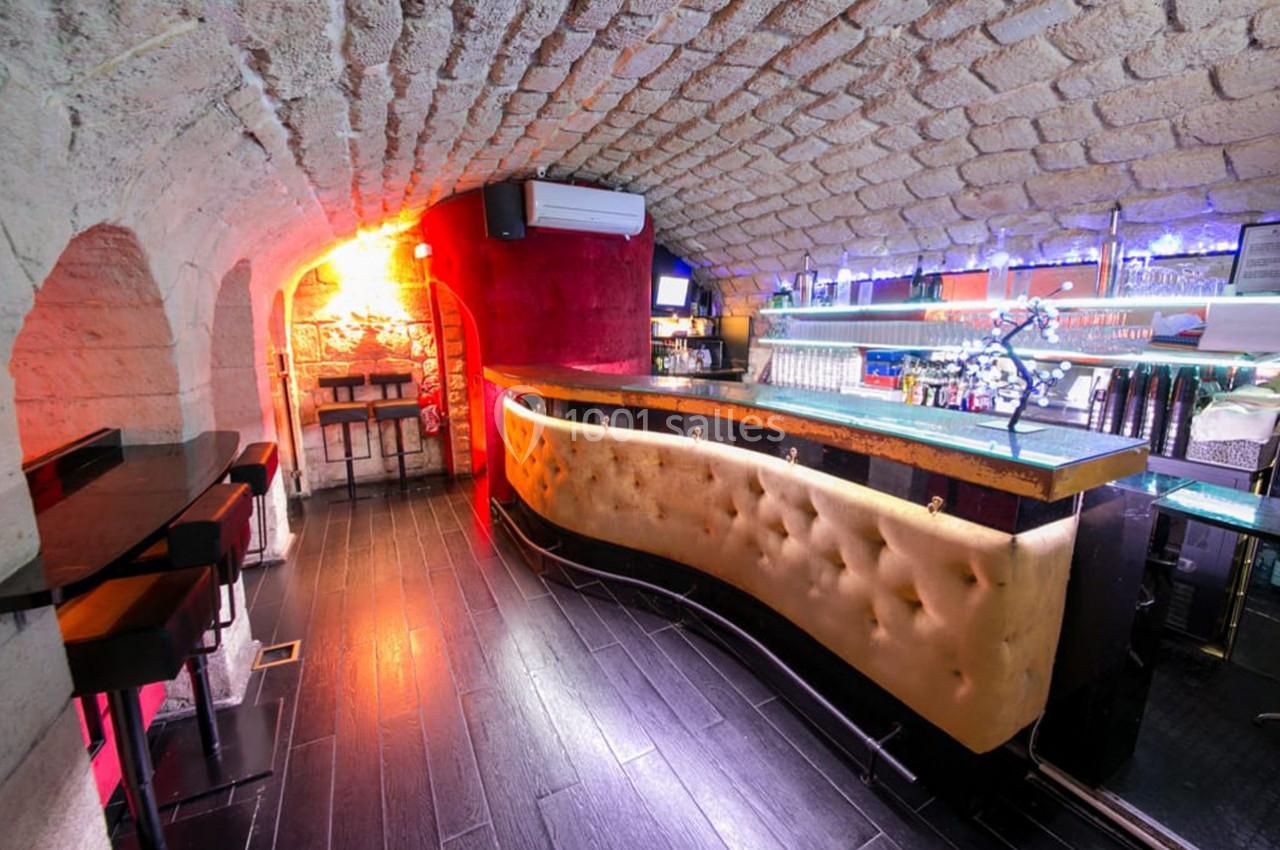 Intérieur d'un bar avec un comptoir capitonné, murs en pierre voûtés et éclairage coloré.