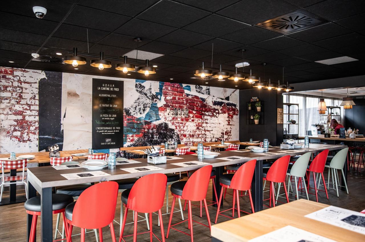 Salle de restaurant moderne avec de longues tables, chaises rouges et mur décoré de briques peintes.