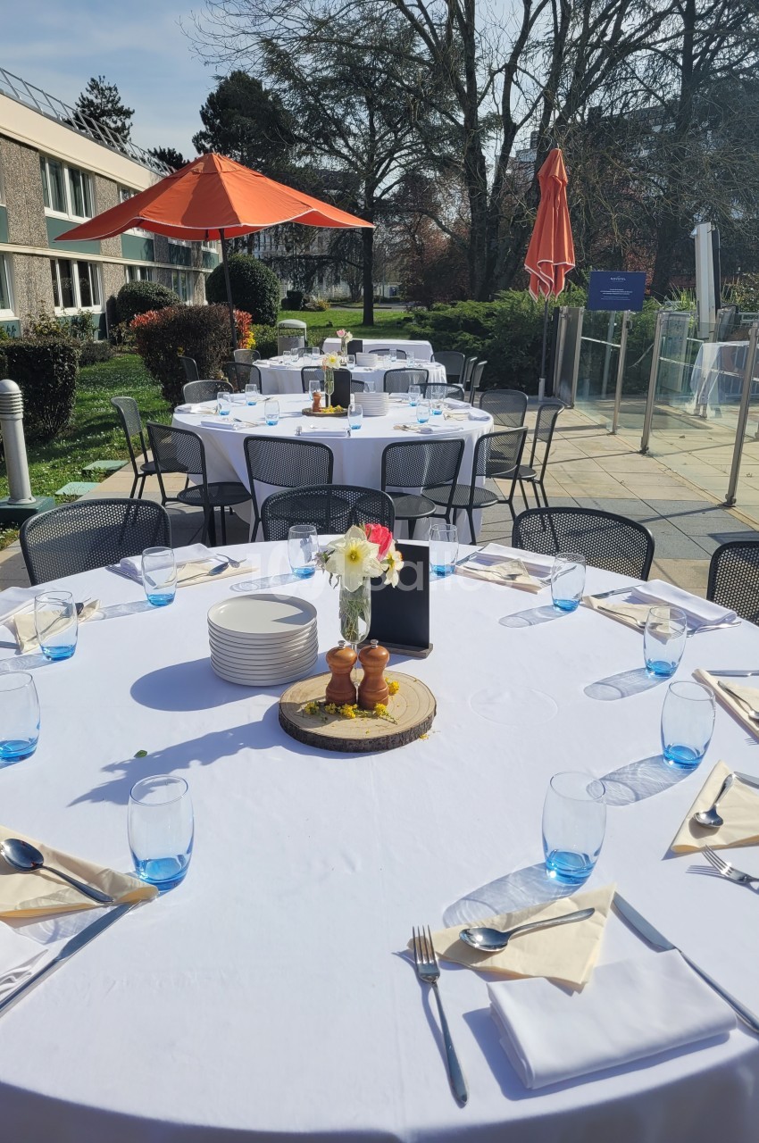 Tables dressées en extérieur avec nappes blanches, verres bleus et décoration florale, sous un ciel ensoleillé.