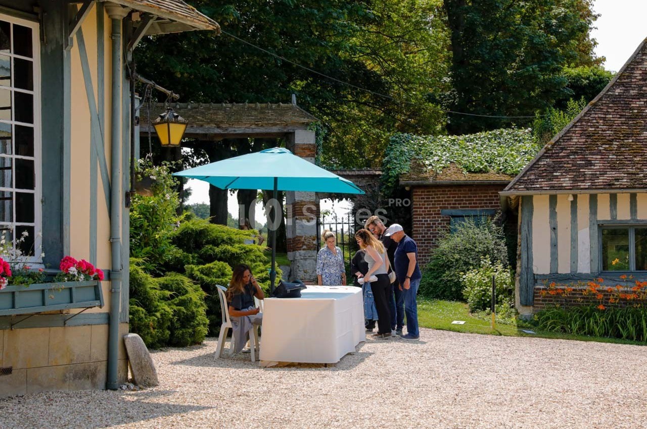 Personnes rassemblées près d'une table sous un parasol bleu dans une cour bordée de bâtiments à colombages.