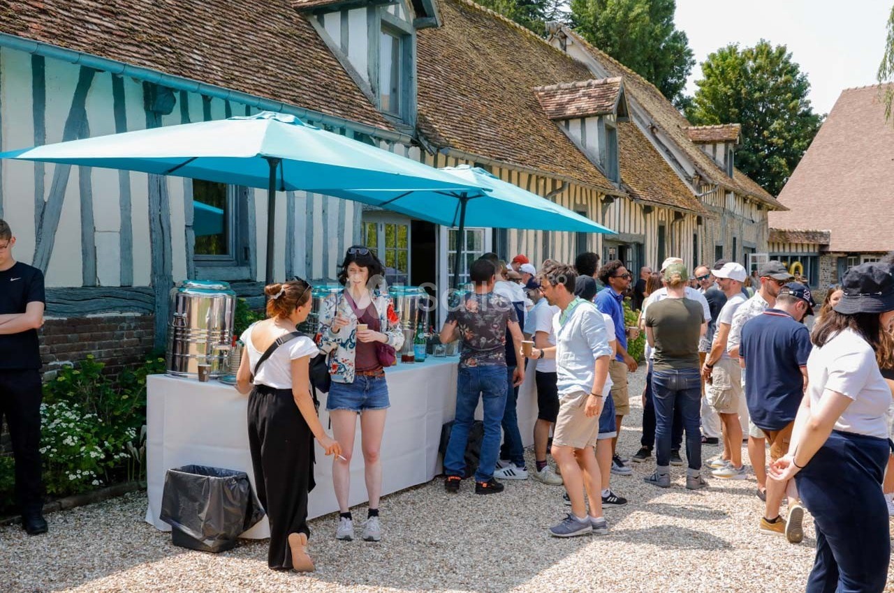 Des personnes font la queue devant un stand de boissons installé en extérieur, près de bâtiments à colombages.