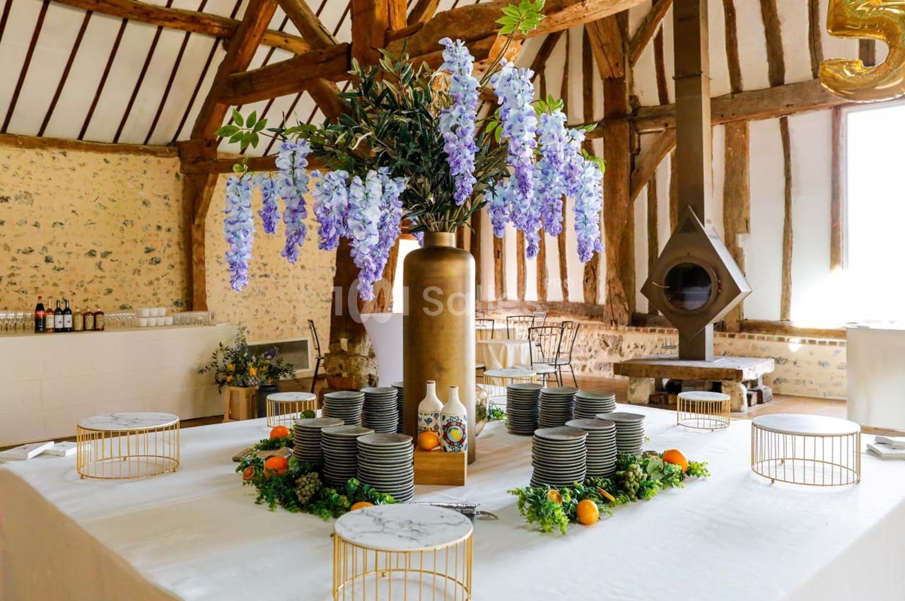 Table décorée avec vaisselle empilée, fleurs violettes dans un vase doré, fruits et verdure, dans une salle rustique.