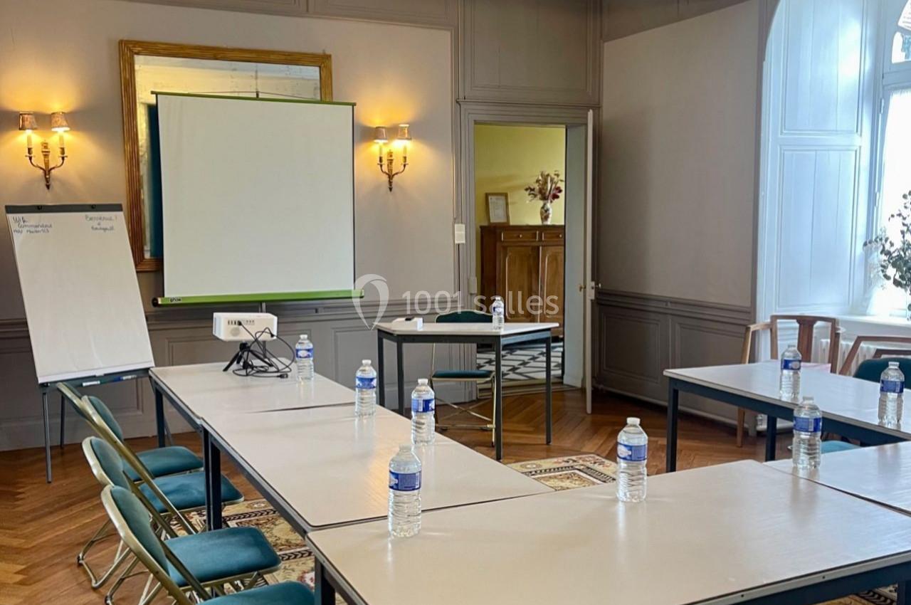 Salle de réunion lumineuse avec tables disposées en U, chaises pliantes, tableau blanc, écran de projection et bouteilles d…