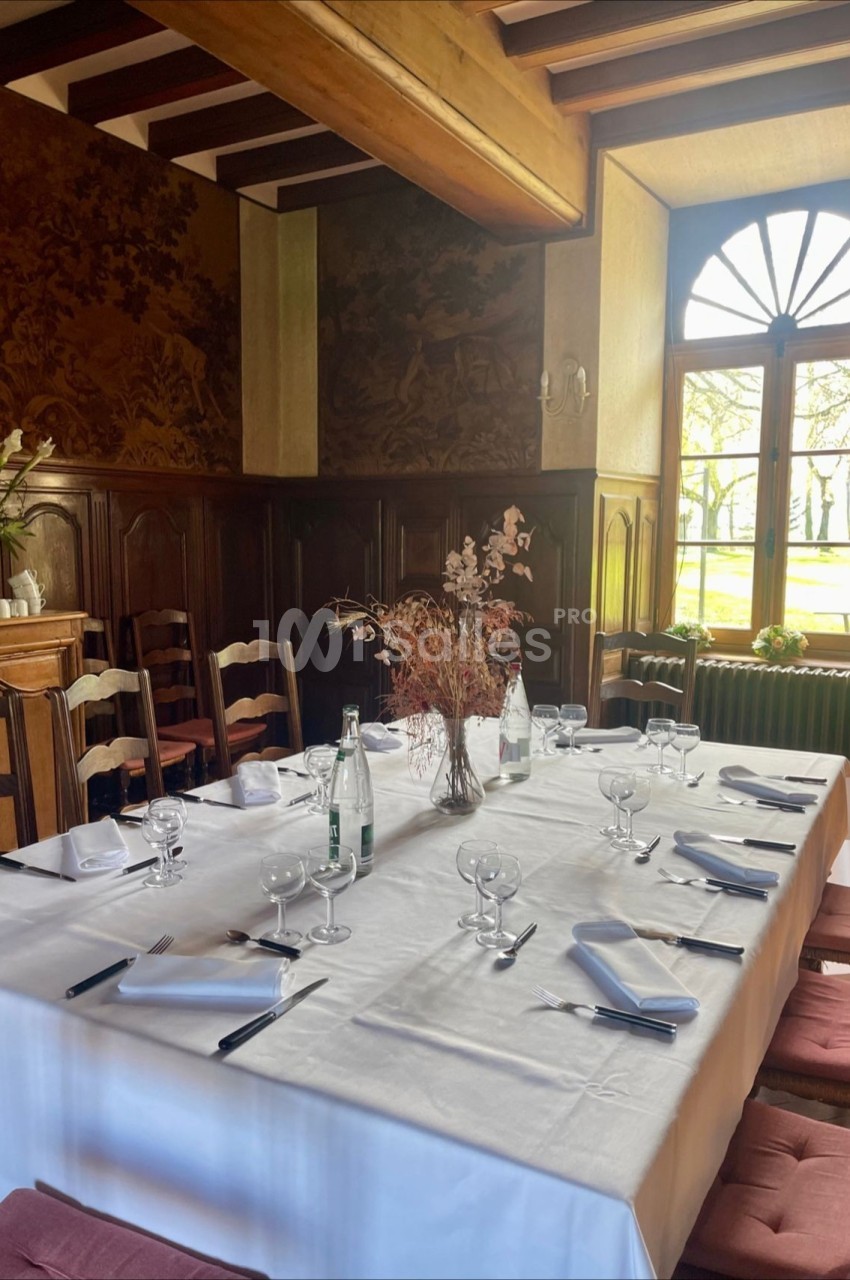 Table dressée avec nappe blanche, verres, couverts et centre de table floral dans une salle lumineuse au décor rustique.