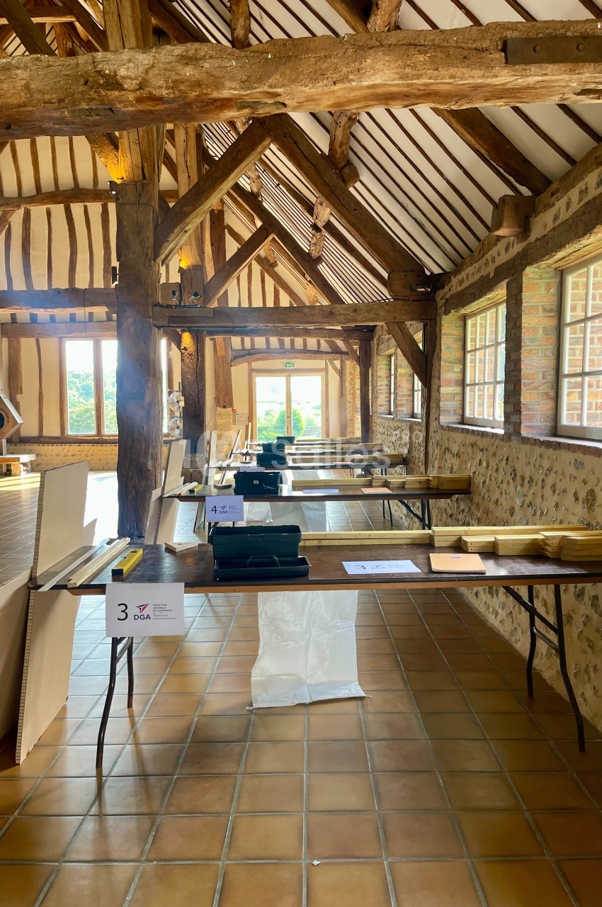 Salle en bois avec des tables alignées, des documents et des équipements, éclairée par des fenêtres et une porte ouverte.