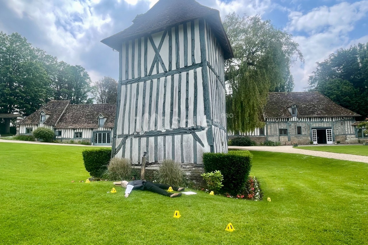 Tour en colombages entourée de pelouse, avec des marqueurs jaunes au sol et une silhouette allongée près de buissons.