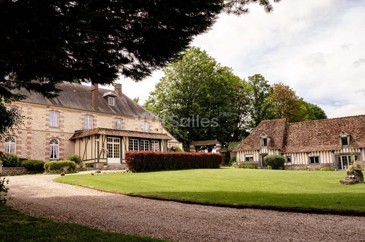 Manoir en pierre et maison à colombages entourés de pelouses, arbres et allée gravillonnée sous un ciel partiellement…