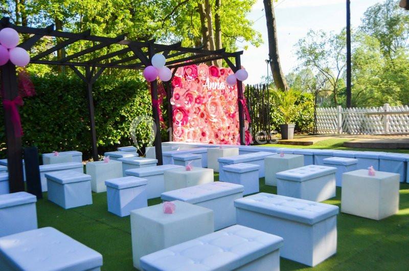 Décor extérieur avec bancs blancs, arche en bois et mur floral rose, entouré de verdure et de clôtures.