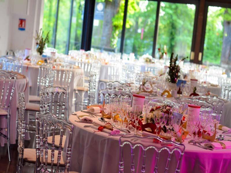 Salle de réception décorée avec des tables rondes, nappes blanches, chaises transparentes et grandes baies vitrées donnant…