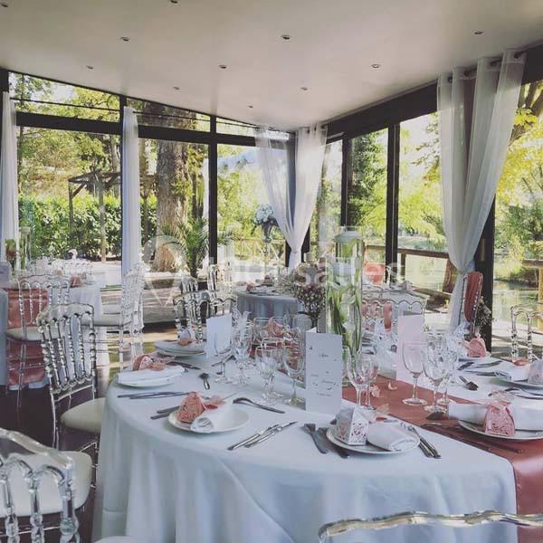 Salle de réception lumineuse avec tables élégamment dressées, nappes blanches et grandes baies vitrées donnant sur un jardin.