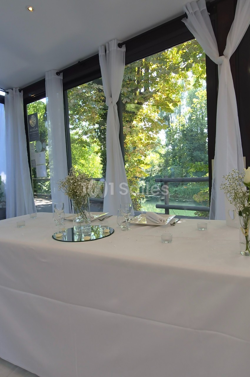 Table décorée avec nappes blanches, fleurs et bougies, placée devant de grandes baies vitrées donnant sur un paysage arboré.