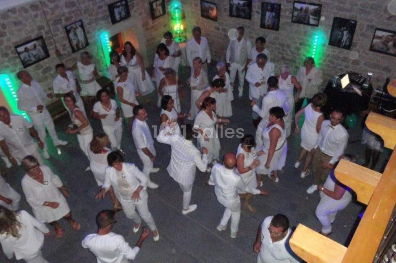 Personnes vêtues de blanc dans une salle en pierre, dansant et socialisant sous des lumières colorées.
