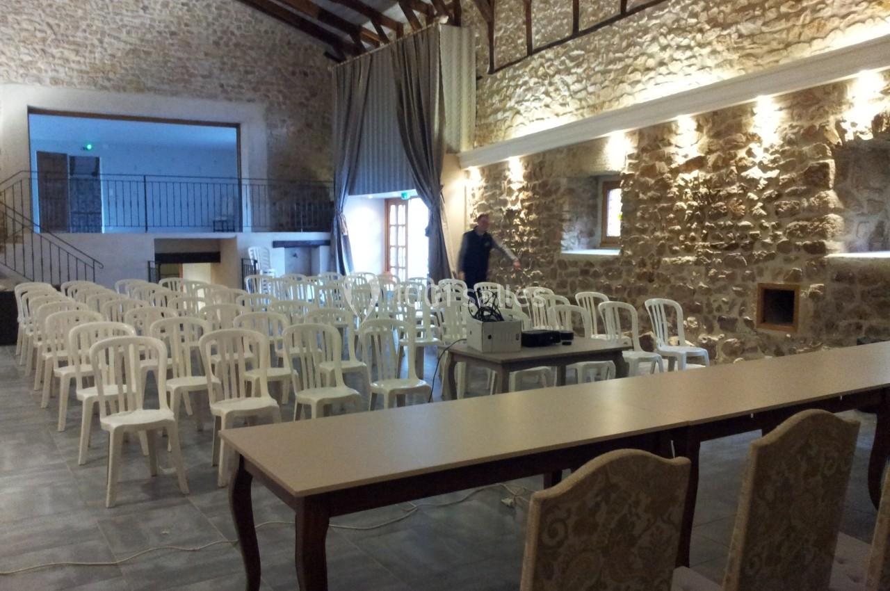 Salle en pierre avec plafond en poutres apparentes, rangées de chaises blanches et tables installées pour un événement.