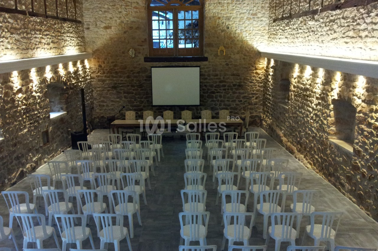 Salle en pierre avec des rangées de chaises blanches disposées face à un écran de projection.