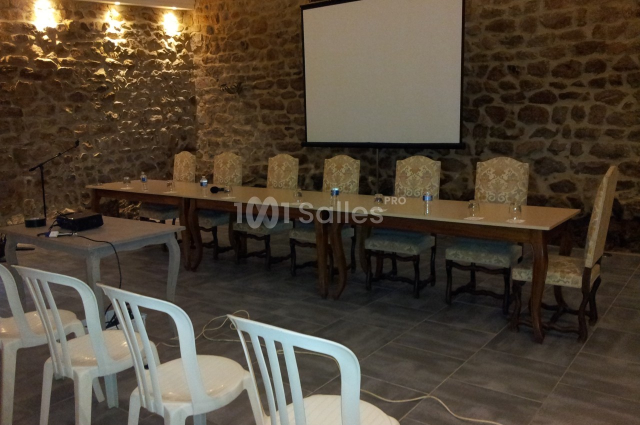 Salle en pierre avec une table longue, chaises alignées, un écran de projection et des chaises en plastique au premier plan.