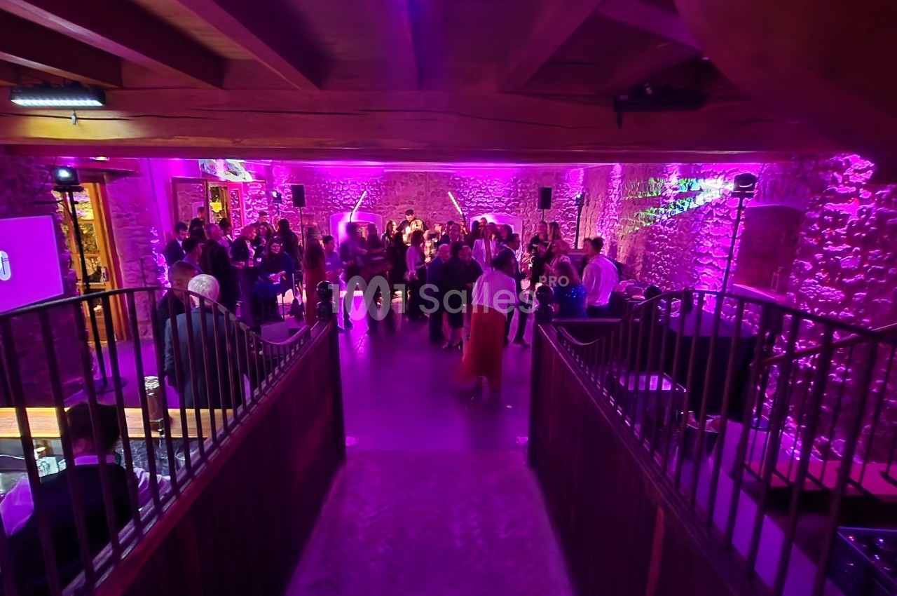 Salle en pierre éclairée de lumières violettes, avec des invités debout ou dansant lors d’un événement festif.