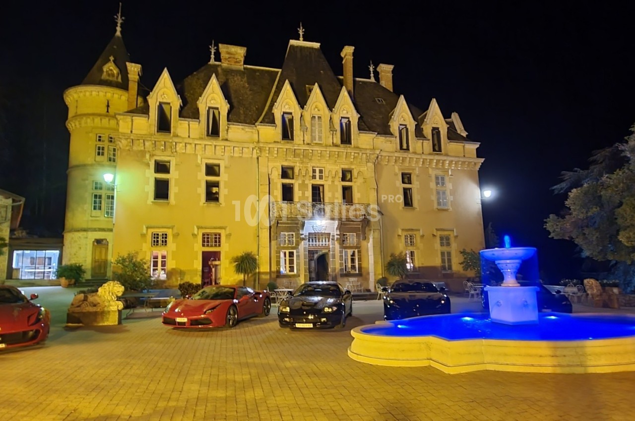 Façade illuminée d'un château de nuit avec une fontaine éclairée en bleu et plusieurs voitures garées devant.