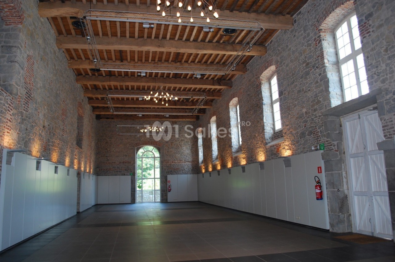Grande salle en pierre avec plafond en bois, fenêtres cintrées, éclairage suspendu et porte vitrée donnant sur l'extérieur.