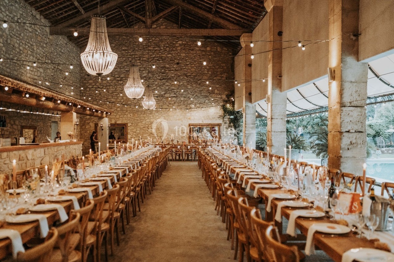 Grande salle en pierre avec de longues tables dressées pour un banquet, éclairée par des lustres et des guirlandes…