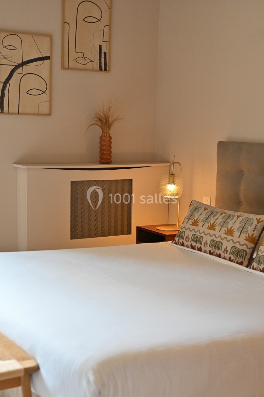 Lit double avec draps blancs, table de chevet avec lampe allumée, décor minimaliste et tableaux abstraits au mur.