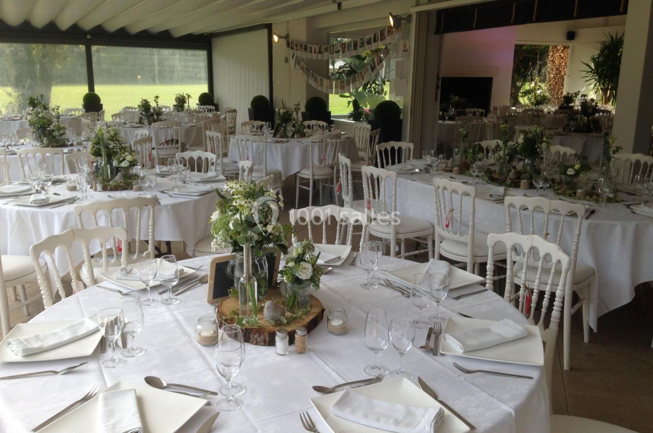 Salle de réception décorée pour un mariage, avec des tables rondes dressées, nappes blanches et arrangements floraux.