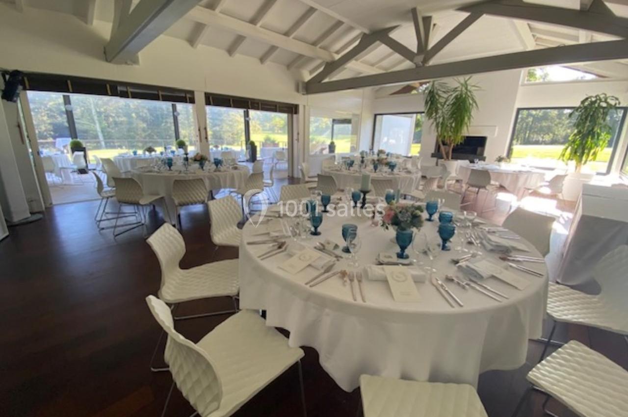 Salle lumineuse avec tables rondes dressées, nappes blanches, verres bleus et vue sur un extérieur verdoyant.