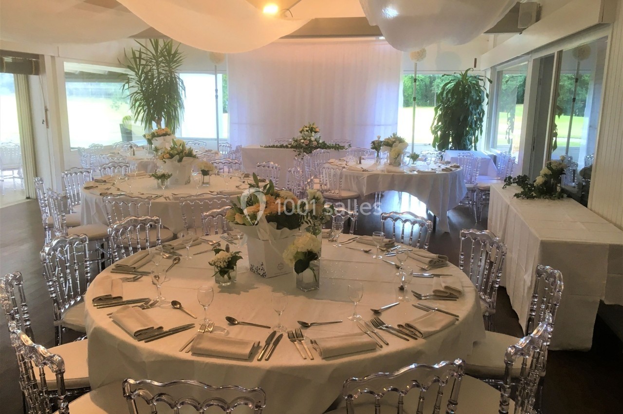 Salle de réception lumineuse avec tables rondes élégamment dressées, décorées de fleurs blanches et jaunes.