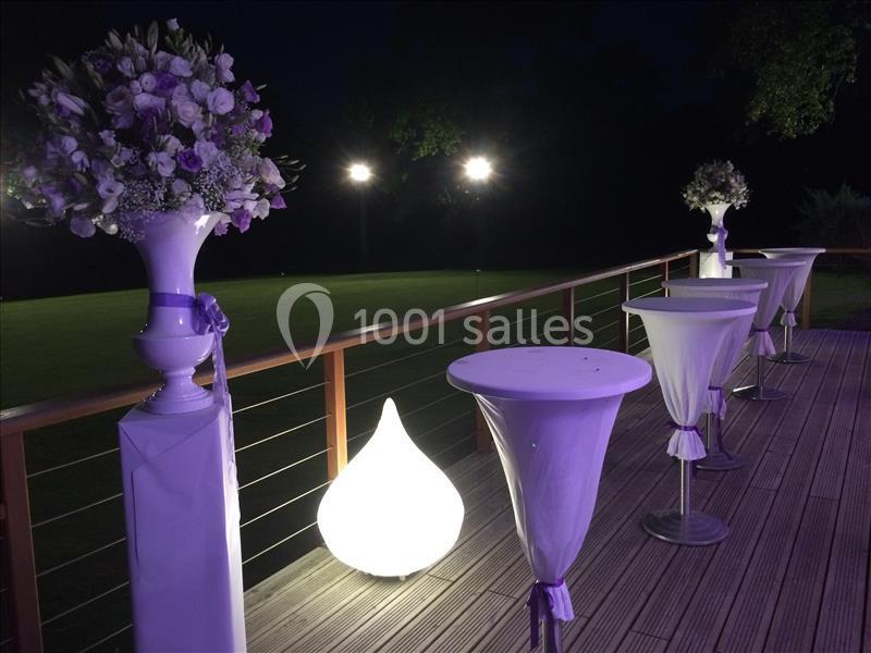 Terrasse éclairée de nuit avec tables hautes drapées et vases de fleurs blanches et violettes.