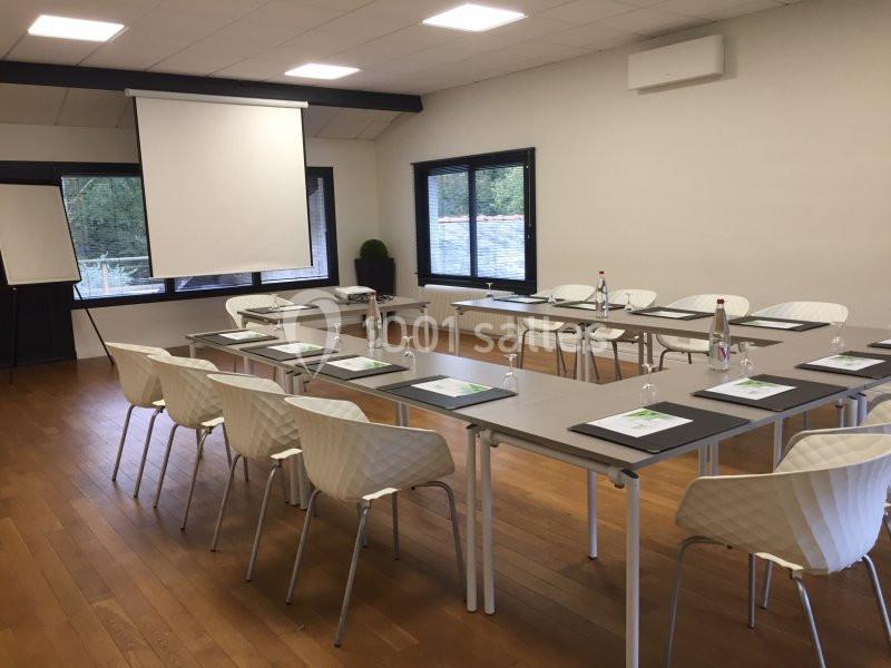 Salle de réunion lumineuse avec tables disposées en U, chaises blanches, écran de projection et paperboard.