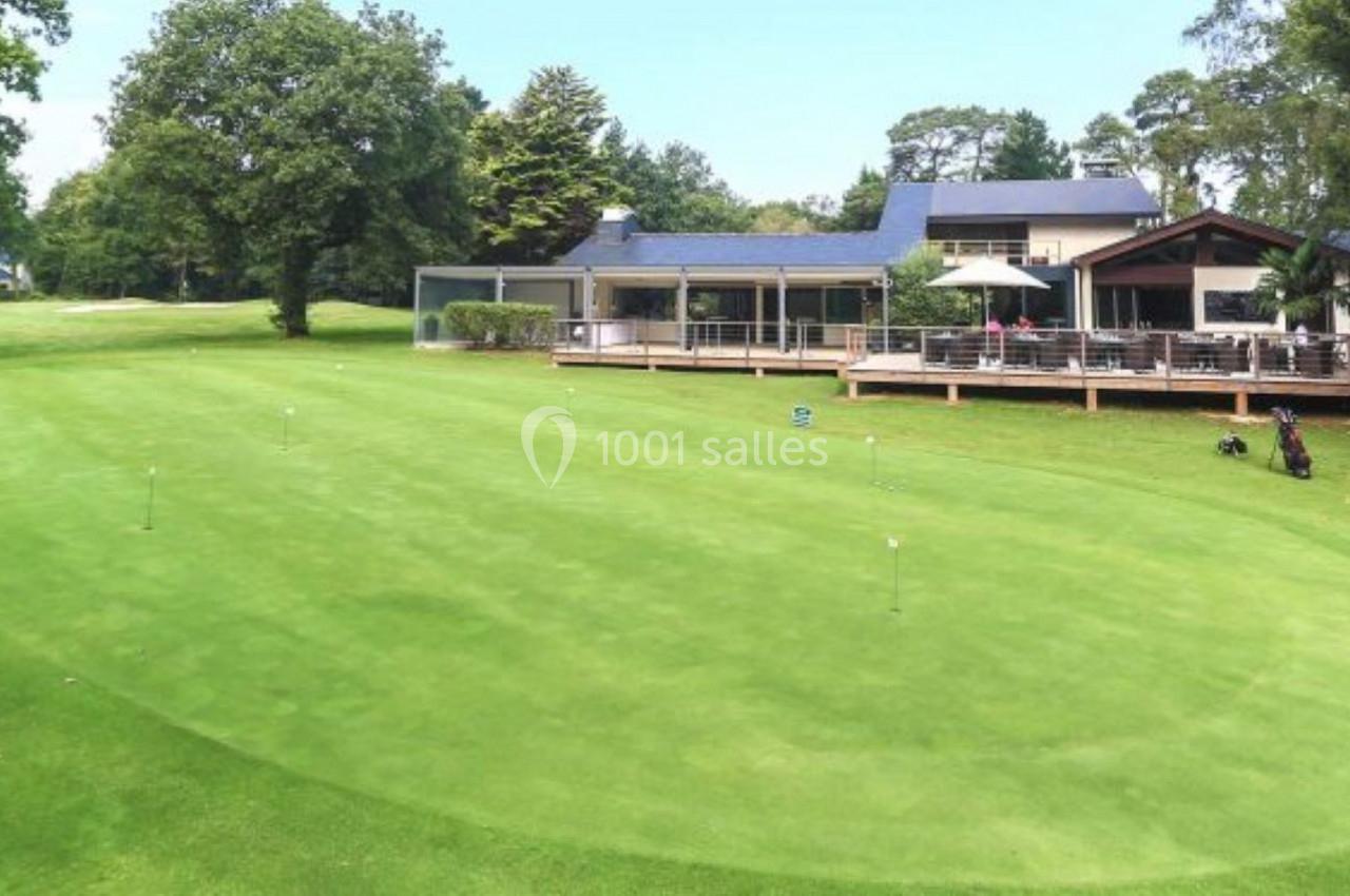 Terrain de golf avec pelouse bien entretenue, entouré d'arbres, et un club-house avec terrasse en arrière-plan.