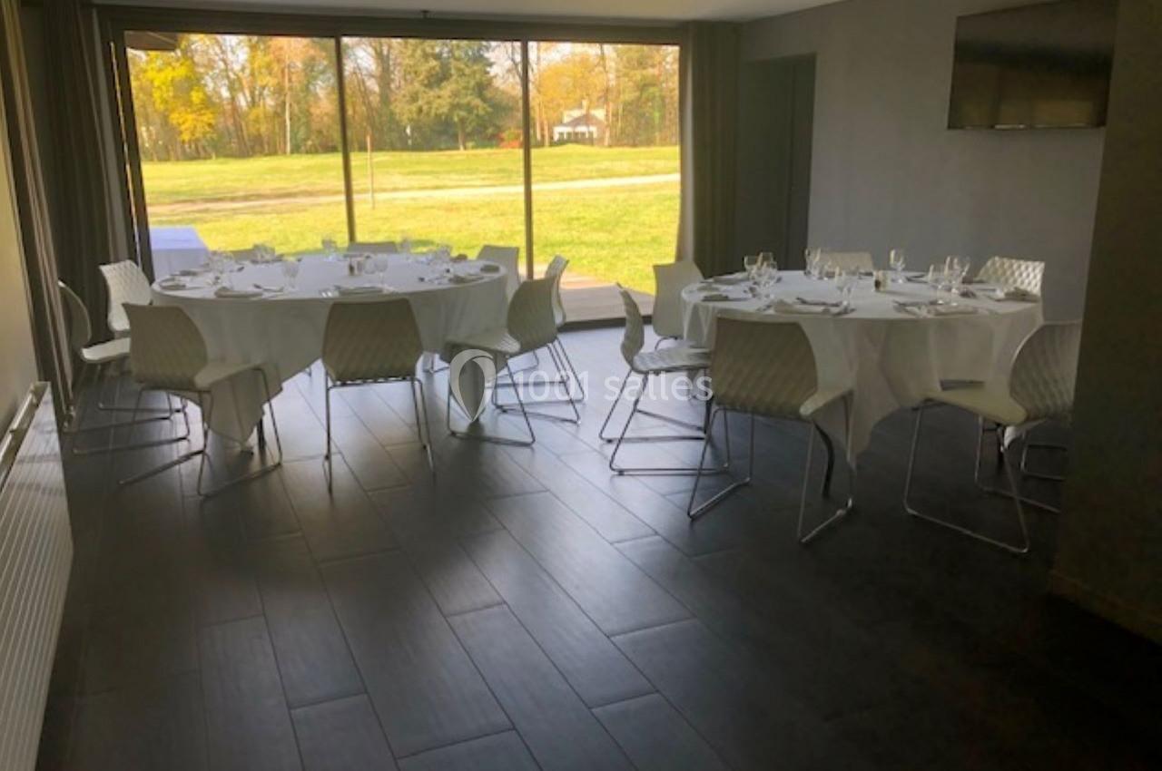 Salle lumineuse avec baies vitrées, tables rondes dressées avec nappes blanches et chaises modernes.