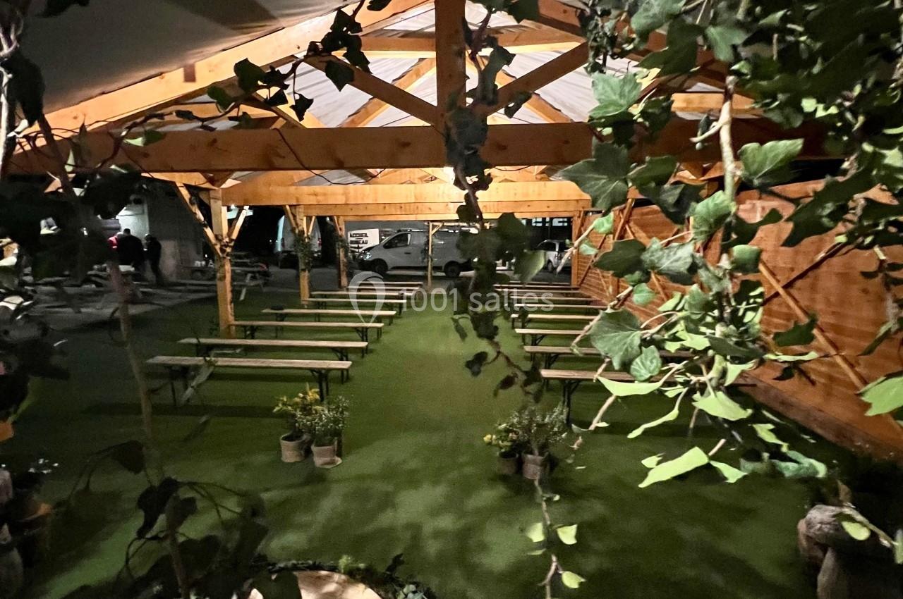 Structure en bois avec bancs alignés sous une tente, entourée de végétation et éclairée par une lumière douce.