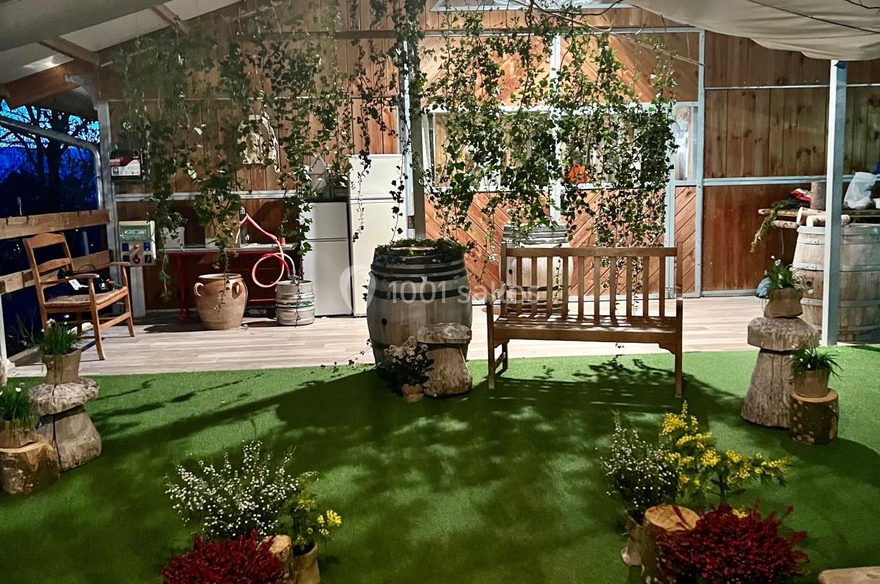 Espace intérieur décoré avec des plantes, tonneaux en bois, banc et sol en gazon artificiel sous une structure en bois.