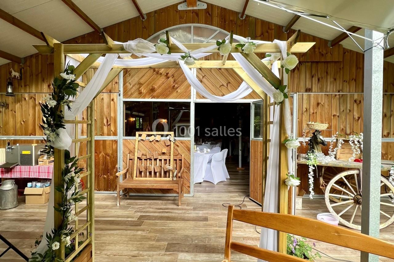Arche décorée de fleurs et de tissu blanc devant une salle en bois aménagée pour un événement festif.