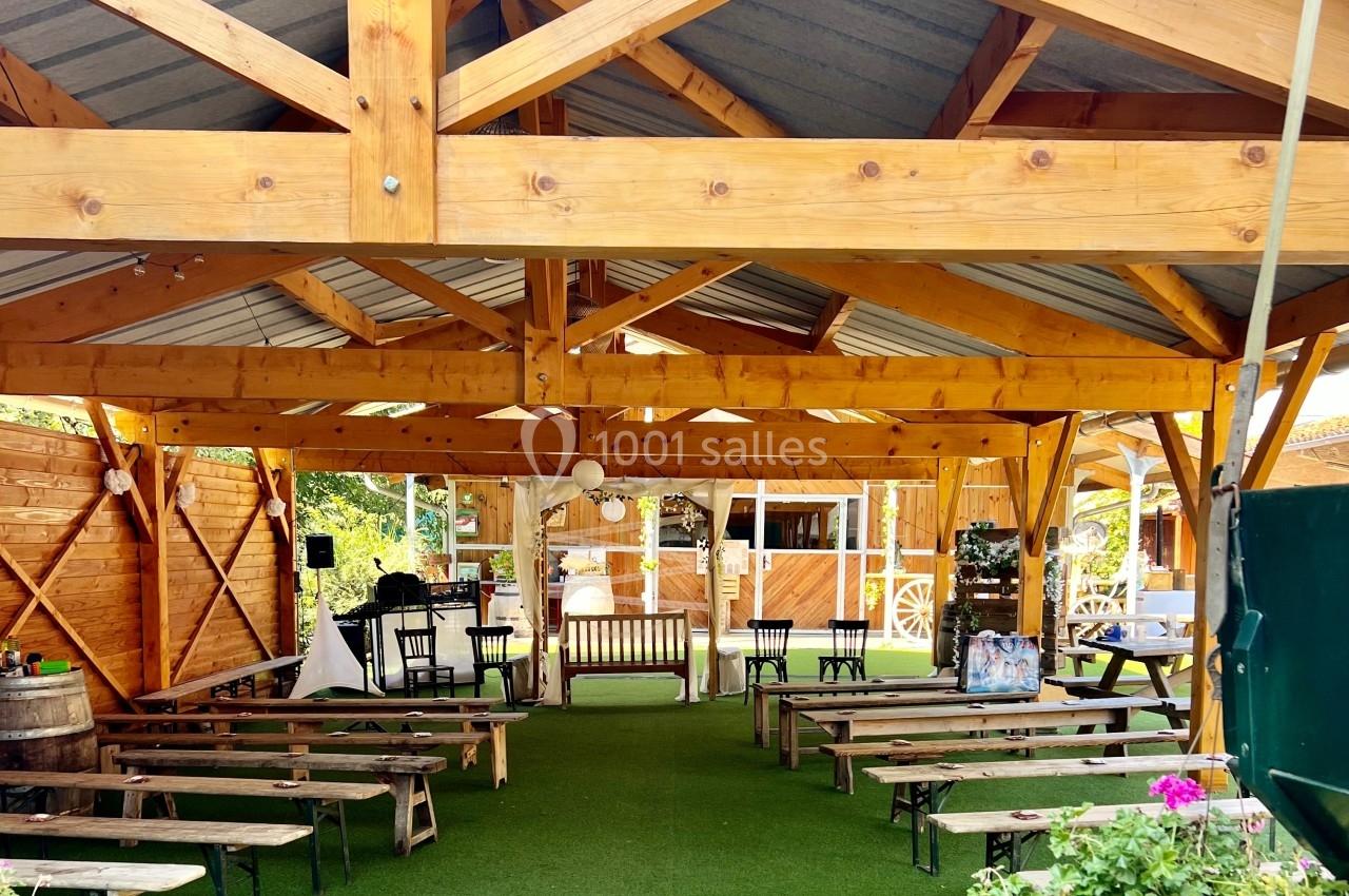 Structure en bois avec toit en métal abritant des bancs et des décorations, entourée de verdure et de fleurs.