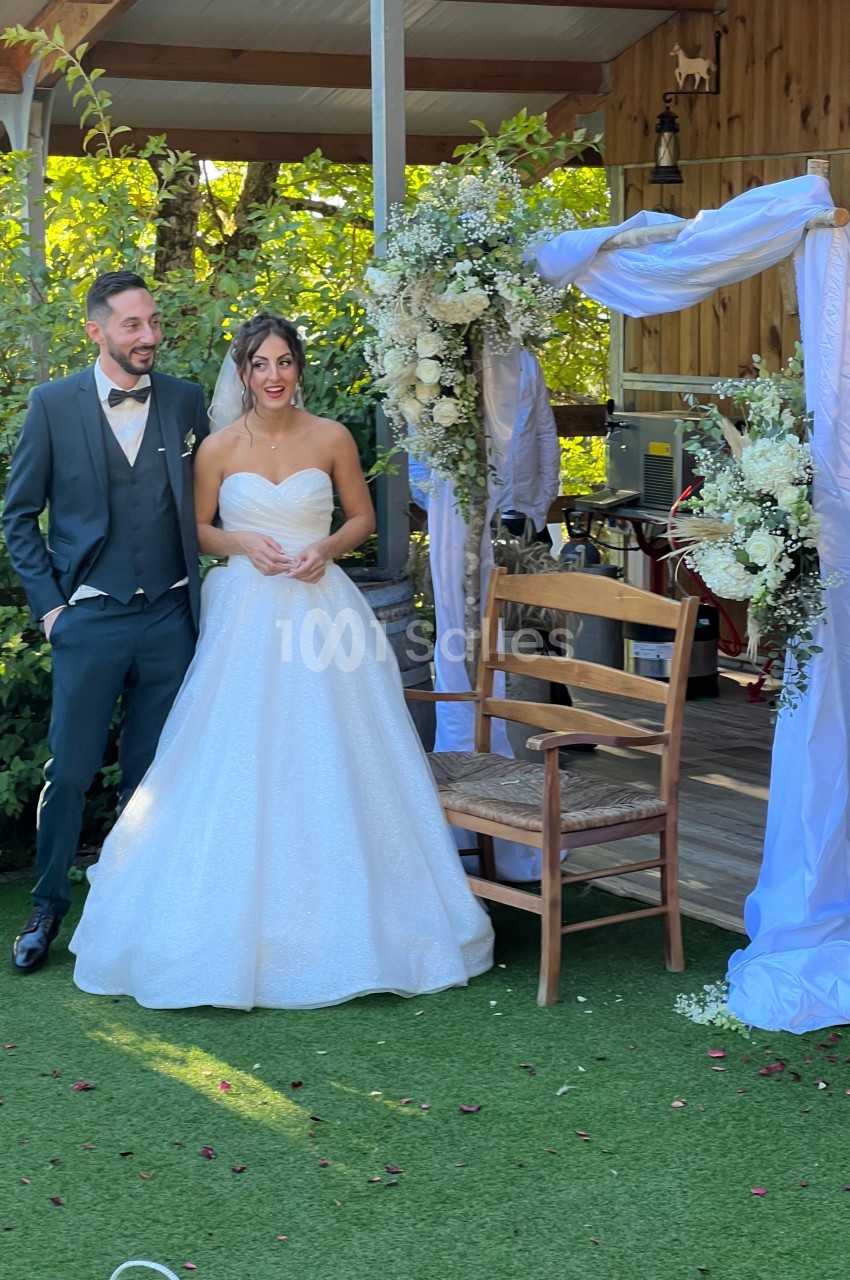 Un couple en tenue de mariage pose près d'une arche décorée de fleurs et de voiles dans un jardin.