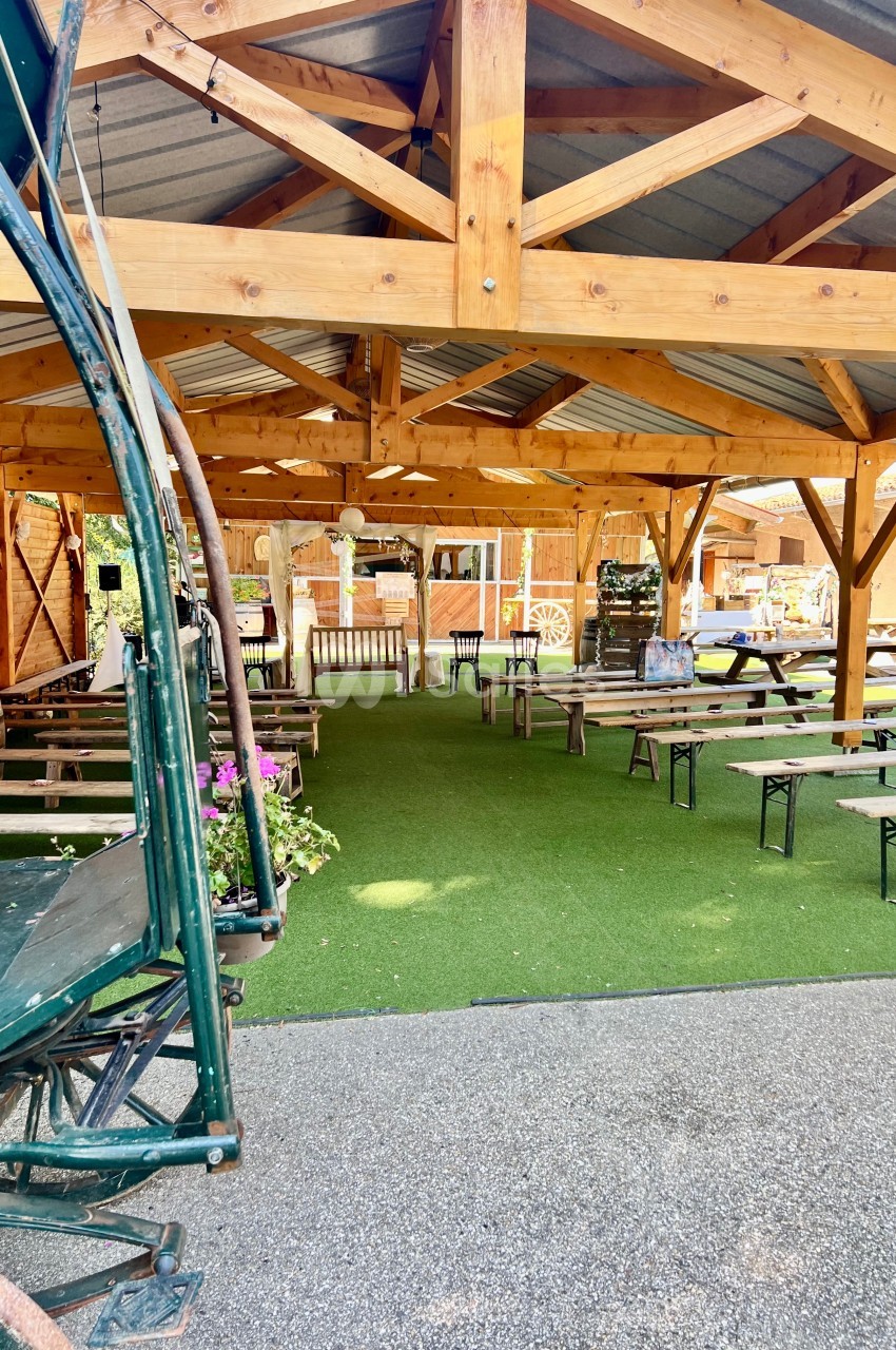 Structure en bois ouverte avec tables et bancs sur un sol en gazon synthétique, dans un espace extérieur aménagé.