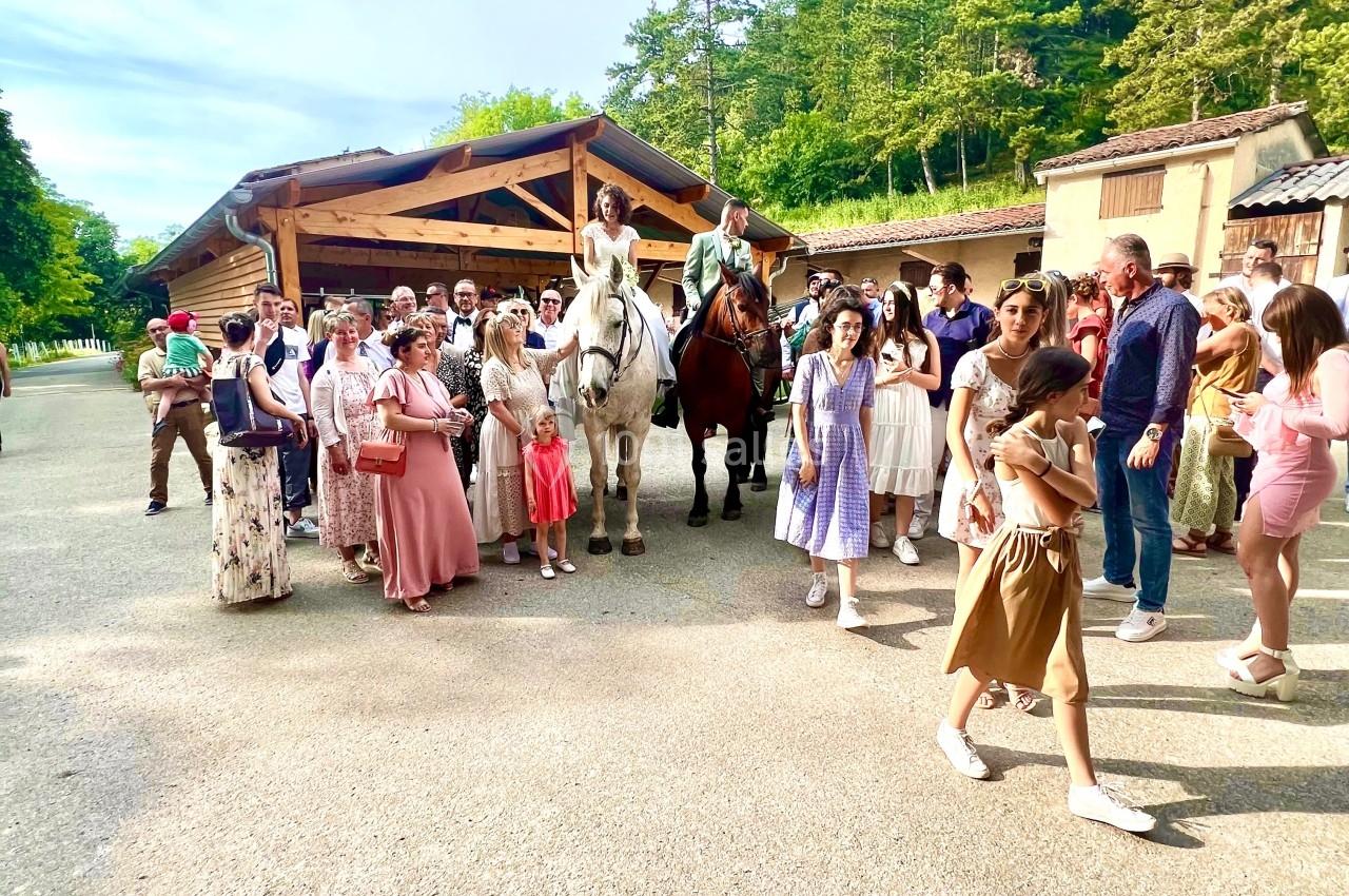 Un groupe de personnes rassemblées en plein air près de chevaux, devant un bâtiment en bois entouré de verdure.