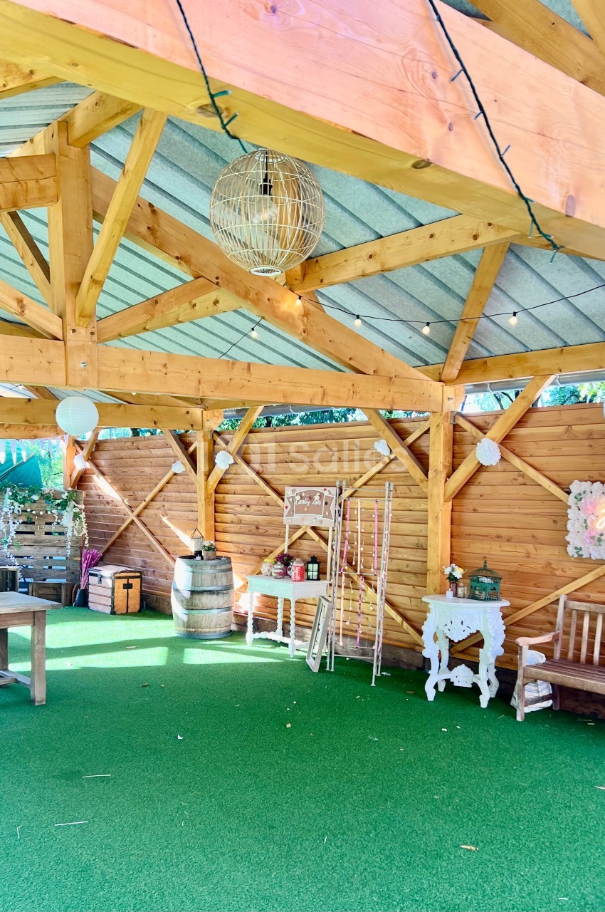 Structure en bois ouverte avec toit métallique, décorée de guirlandes lumineuses, mobilier rustique et fleurs.