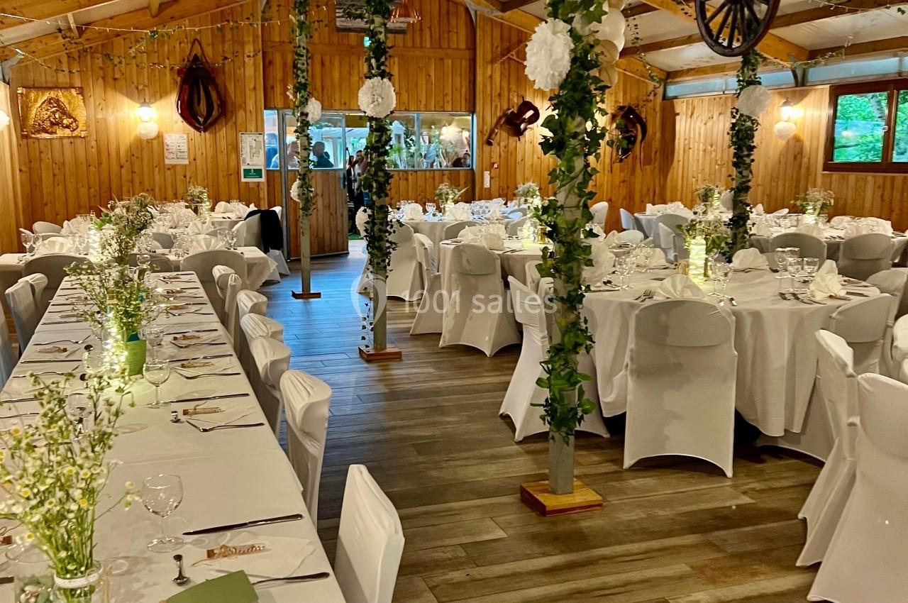 Salle de réception en bois décorée avec des tables dressées, des chaises blanches et des guirlandes végétales.