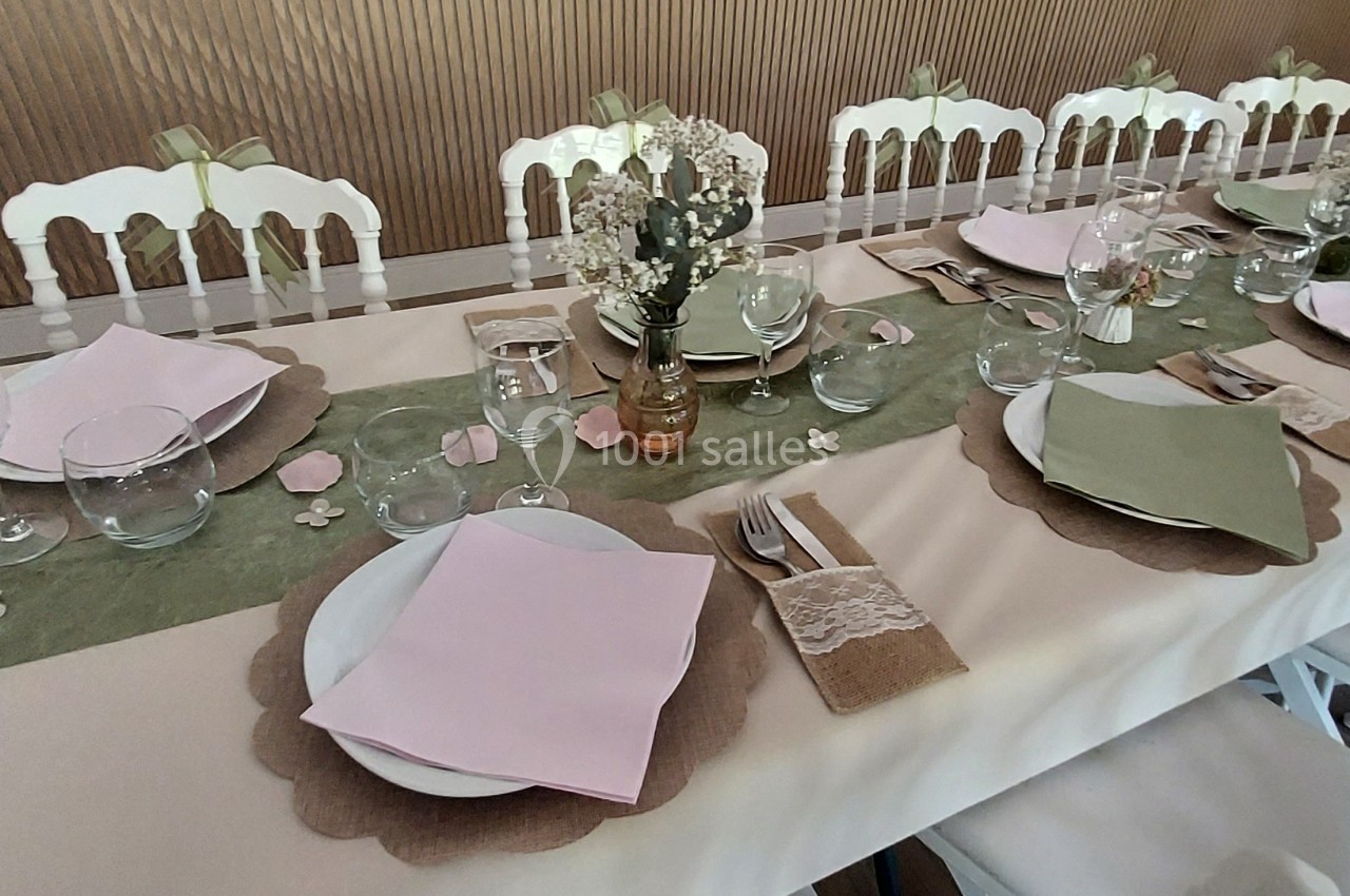 Table décorée pour un événement avec nappes beiges, serviettes roses et vertes, vaisselle élégante et centre de table fleuri.