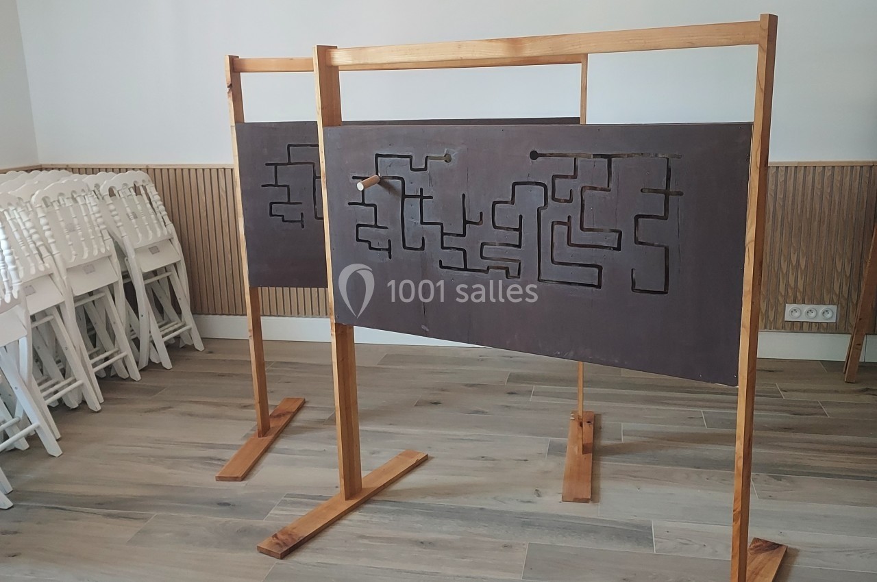 Panneau en bois avec un labyrinthe gravé, monté sur un support, dans une pièce avec des chaises empilées.