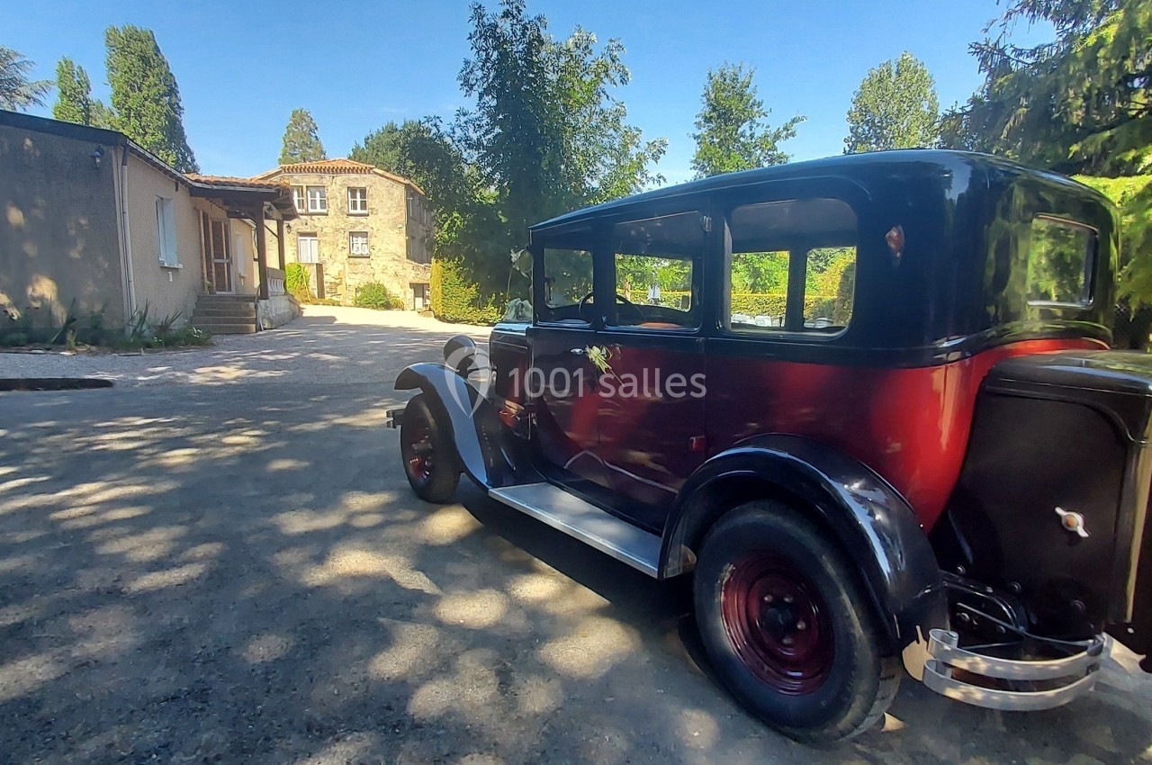 Voiture ancienne rouge et noire stationnée dans une cour pavée entourée de bâtiments en pierre et de végétation.