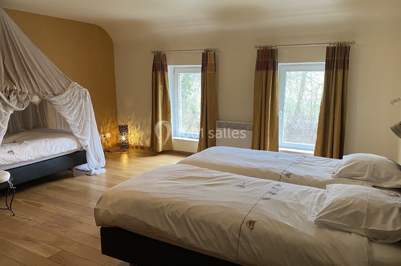 Chambre lumineuse avec deux lits simples, un lit à baldaquin, parquet en bois et fenêtres donnant sur un extérieur arboré.