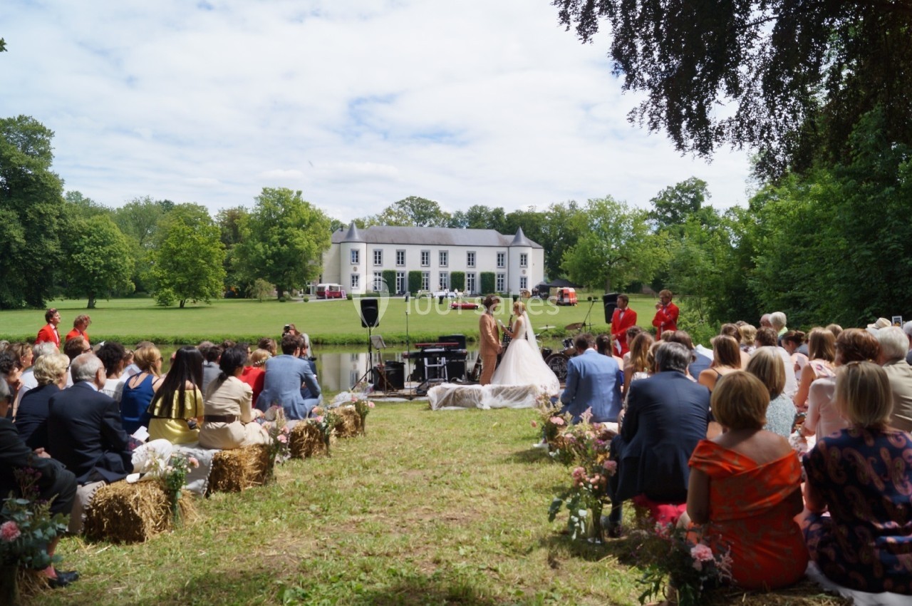 Cérémonie de mariage en plein air devant un manoir, avec des invités assis et des mariés près d'un officiant.