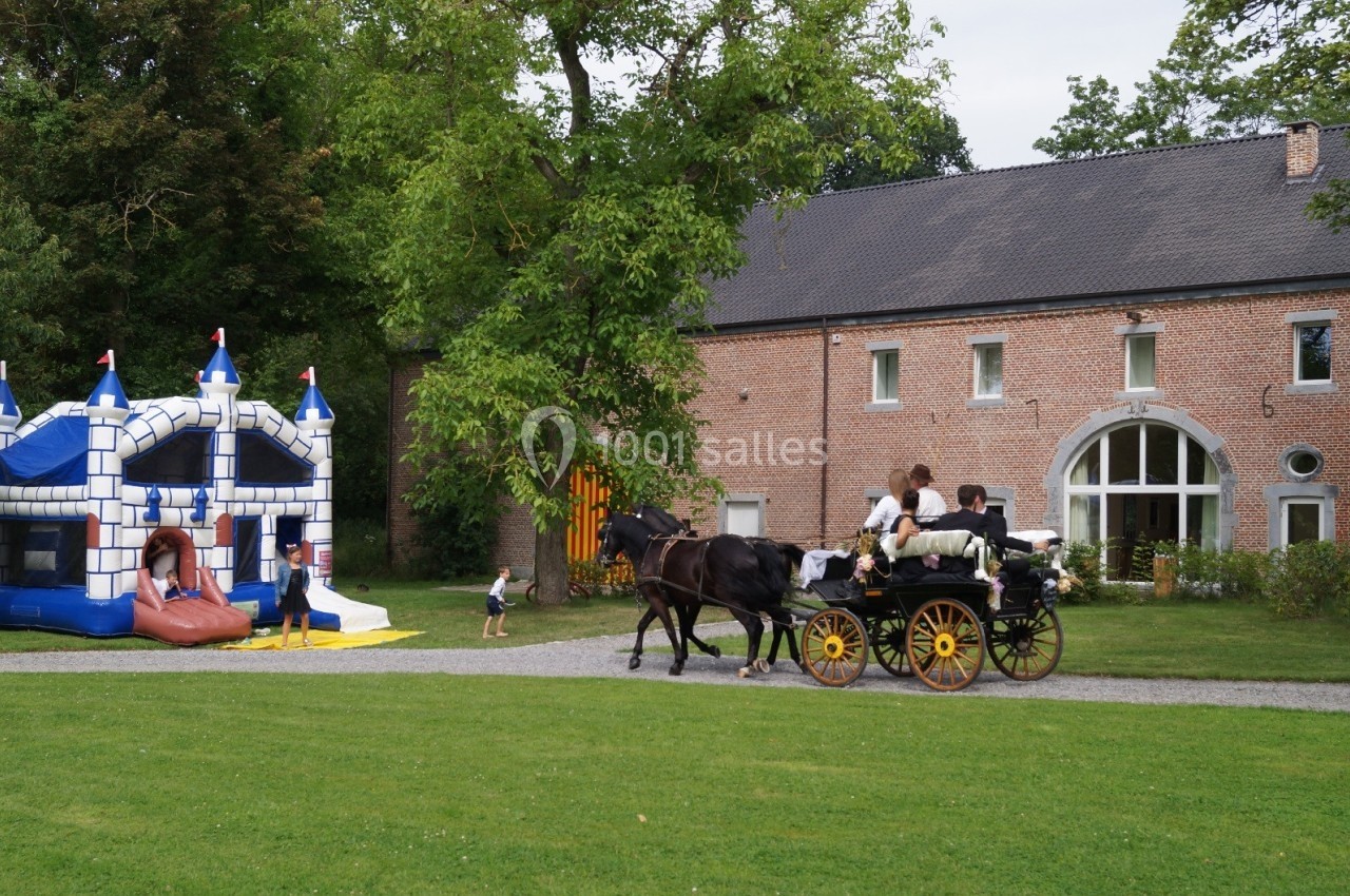 Une calèche tirée par deux chevaux passe devant une maison en briques, près d'un château gonflable dans un jardin.