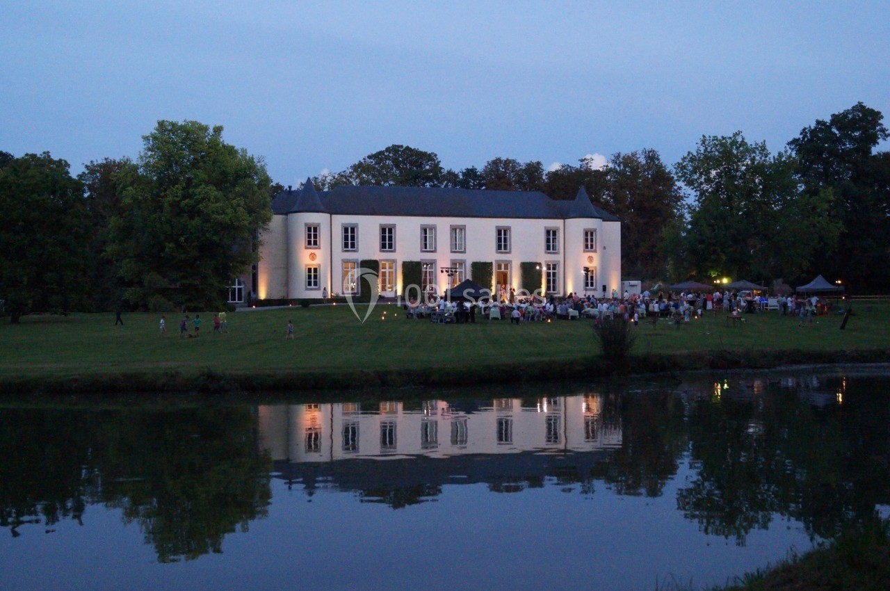 Manoir éclairé au crépuscule avec un rassemblement de personnes sur une pelouse, reflet visible dans un étang.