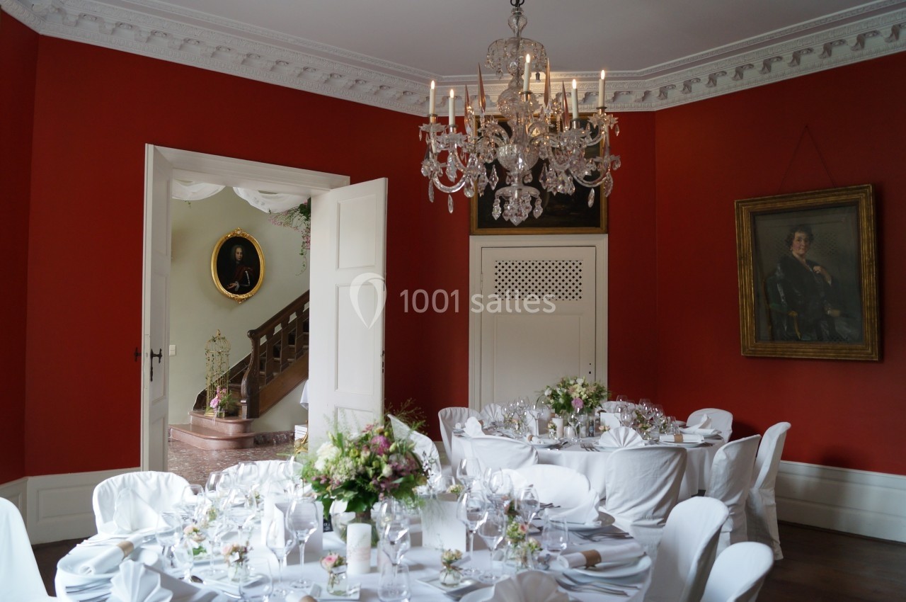 Salle à manger élégante avec murs rouges, tables dressées pour un événement, lustre en cristal et tableaux accrochés.